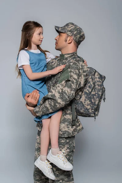 Soldado del ejército en uniforme militar y mochila abrazando hija sobre fondo gris - foto de stock