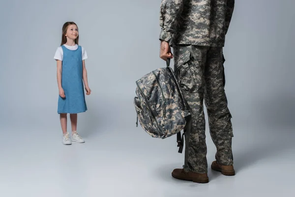 Glückliches Mädchen blickt Vater in Militäruniform mit Rucksack bei der Heimkehr auf grauem Hintergrund an — Stockfoto