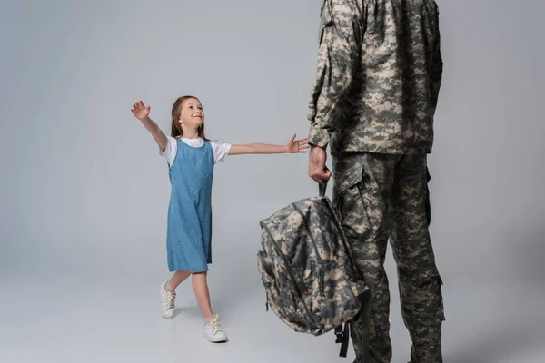 Fille heureuse accueillant père en uniforme militaire avec sac à dos pendant le retour à la maison sur fond gris — Photo de stock