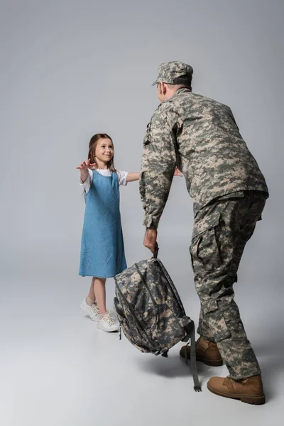 Happy kid welcoming father in military uniform with backpack during homecoming on grey background — Stock Photo