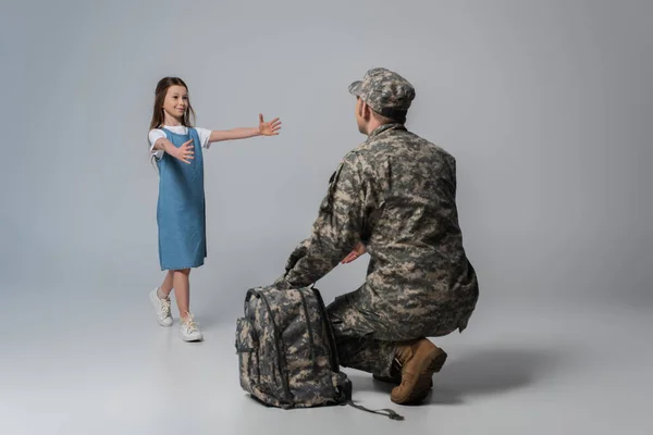 Щаслива дитина вітає батька в армійській формі під час перегляду на сірому фоні — стокове фото