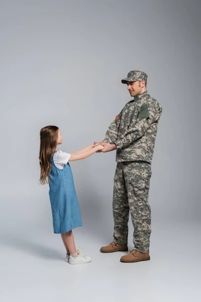 Full length of happy serviceman in army uniform holding hands with daughter on grey — Stock Photo