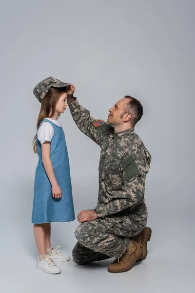 Feliz serviceman em uniforme do exército vestindo tampão militar na cabeça da filha bonito no cinza — Fotografia de Stock