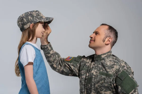 Веселый военнослужащий в военной форме в военной фуражке на голове дочери во время памятного дня, изолированного по серому — стоковое фото