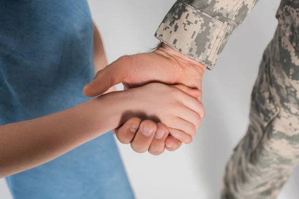 Vista recortada de niña preadolescente cogida de la mano del valiente padre en uniforme del ejército durante el día conmemorativo aislado en gris - foto de stock