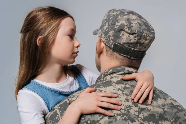 Chica patriótica mirando al padre en uniforme del ejército y llorando durante el día conmemorativo aislado en gris - foto de stock