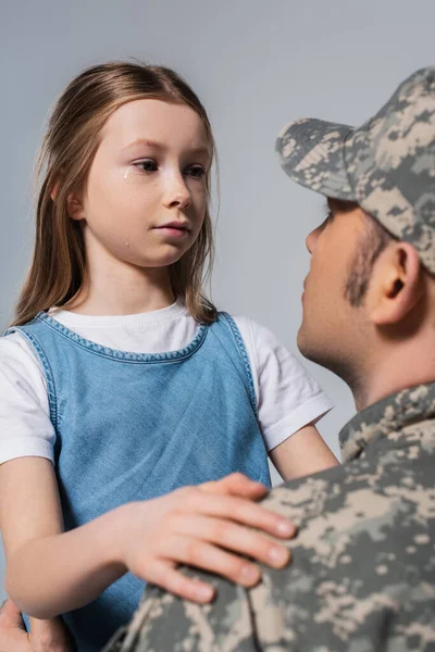 Chica patriótica mirando al militar en uniforme del ejército y llorando durante el día conmemorativo aislado en gris - foto de stock
