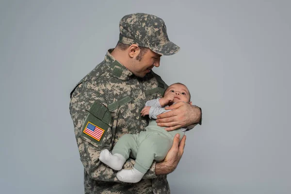Щасливий солдат у формі і шапка тримає новонародженого сина ізольовано на сірому — стокове фото