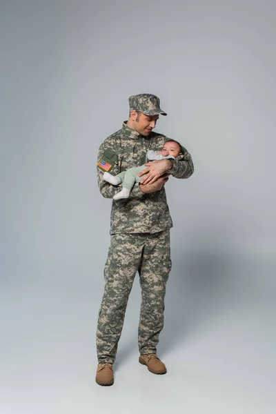 Full length of cheerful serviceman in uniform and cap holding newborn son on grey — Stock Photo