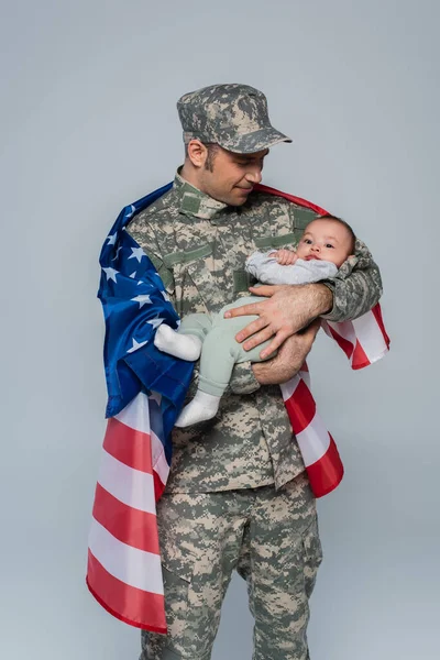 Patriotic serviceman in uniform and cap standing with American flag and holding newborn son isolated on grey — Stock Photo