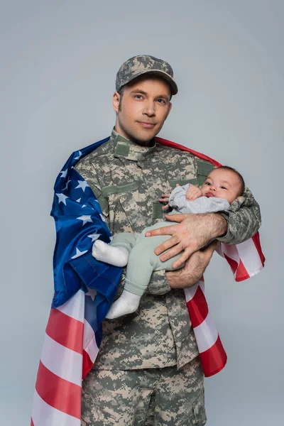 Militar patriótico en uniforme de pie con la bandera americana y la celebración de hijo recién nacido aislado en gris - foto de stock