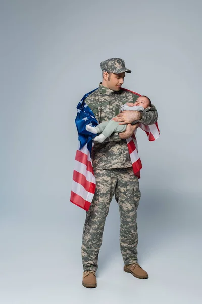 Militar patriótico en uniforme y gorra de pie con bandera americana y sosteniendo al niño en brazos sobre gris - foto de stock