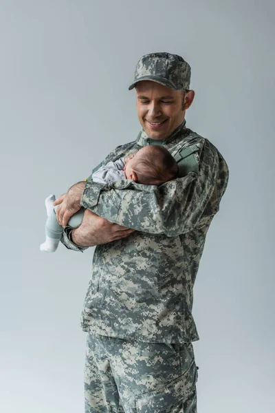 Cheerful serviceman in uniform and cap holding newborn son isolated on grey — Stock Photo