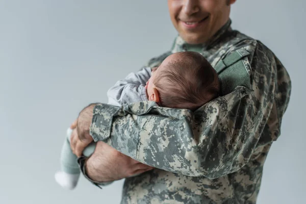 Cheerful serviceman in camouflage uniform holding newborn son isolated on grey — Stock Photo