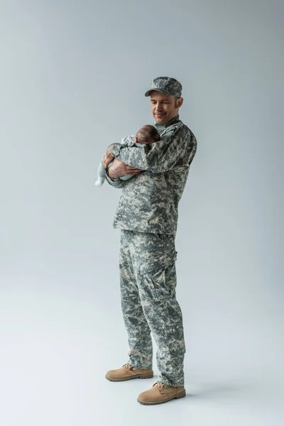 Full length of army serviceman in uniform and cap holding newborn son on grey — Stock Photo