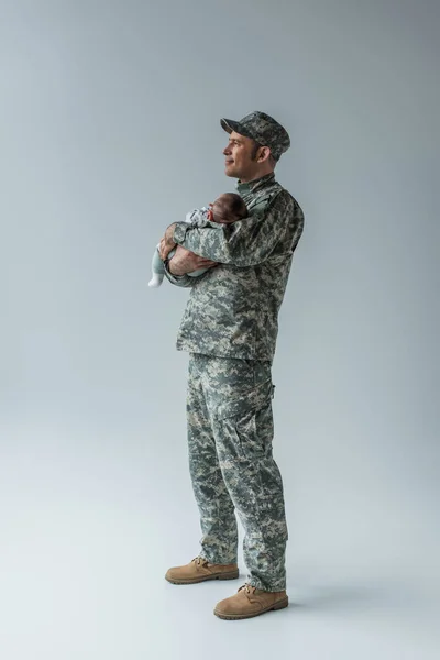 Повна довжина приємного військовослужбовця в формі і кепка тримає новонародженого сина на сірому — стокове фото