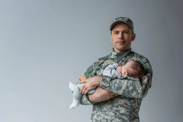 Militante americano en uniforme y gorra sujetando hijo recién nacido aislado en gris - foto de stock