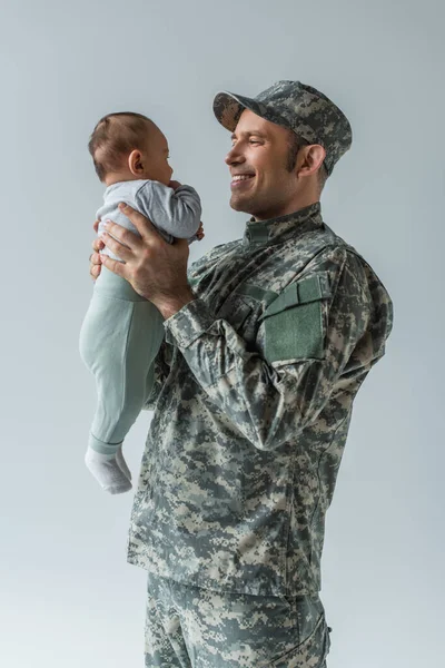 Солдат в форме, держащий новорожденного сына в руках, изолированного на сером — стоковое фото