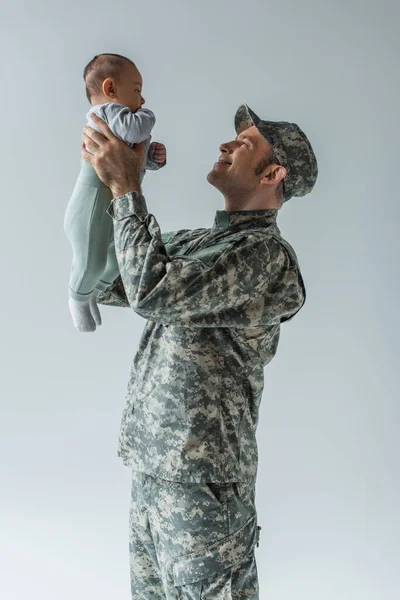Soldado del ejército feliz en uniforme sosteniendo bebé niño en brazos aislado en gris - foto de stock