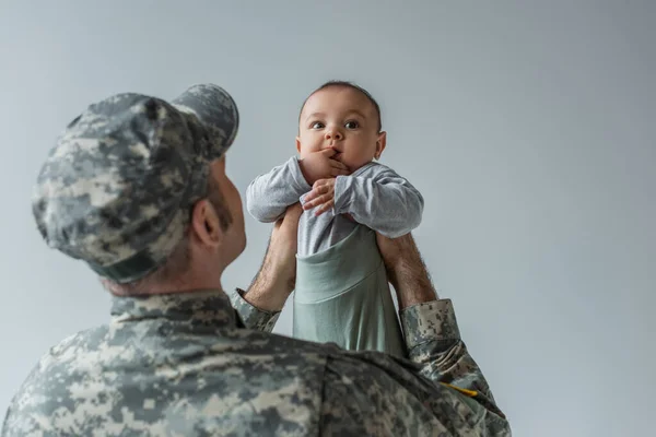 Армейский солдат в военной форме и кепке, держащий маленького сына на руках изолированным на сером — стоковое фото