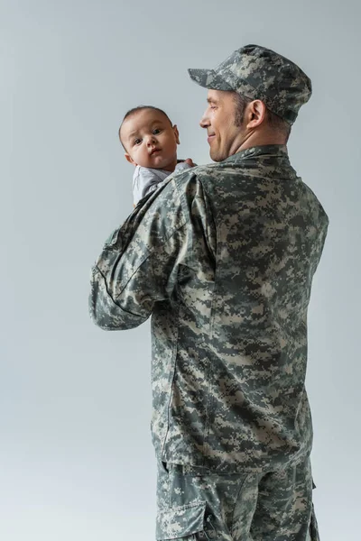 Улыбающийся армейский солдат в военной форме с шапкой, держащий младенца в руках, изолированного на сером — стоковое фото