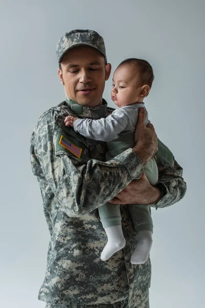 Soldado do exército americano em uniforme militar com boné segurando filho bebê em armas isoladas em cinza — Fotografia de Stock