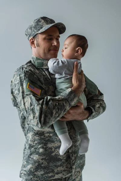Soldado del ejército estadounidense en uniforme militar sosteniendo a su hijo en armas aislado en gris - foto de stock