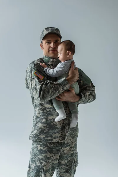 Довольный американский военнослужащий в военной форме, держащий в руках новорожденного сына, изолированного на сером — стоковое фото