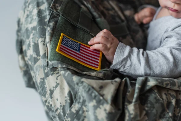 Обрезанный вид американского солдата в форме, держащего в руках новорожденного мальчика, изолированного на сером — стоковое фото