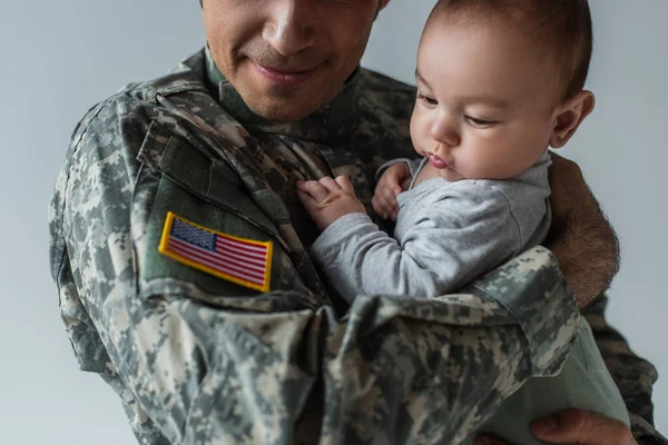 Американський солдат, який дивиться на прапор на формі, тримаючи в руках новонародженого хлопчика, ізольованого на сірому — стокове фото
