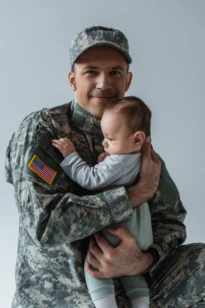 Soldado estadounidense feliz en uniforme sosteniendo en brazos niño recién nacido aislado en gris - foto de stock