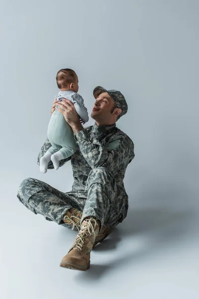 Солдат в форме, держащий в руках новорожденного мальчика, сидящего на сером — стоковое фото