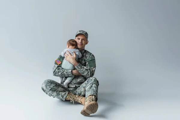 USA soldier in uniform holding in arms newborn boy while sitting on grey — Stock Photo