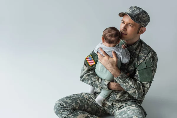 Гордый отец в военной форме, держащий в руках новорожденного сына, сидящего на сером фоне — стоковое фото