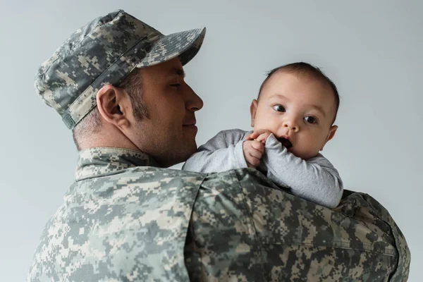 Весёлый отец в военной форме и кепке обнимает новорожденного сына, изолированного на сером — стоковое фото