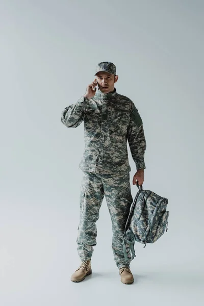 Serviceman in cap and army uniform talking on smartphone while holding backpack on grey — Stock Photo