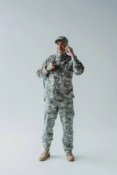 Full length of happy serviceman in cap and uniform talking on smartphone on grey — Stock Photo