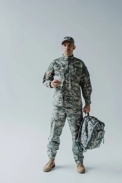 Soldado americano en uniforme del ejército con bandera nacional sosteniendo smartphone y mochila en gris - foto de stock