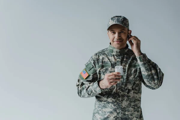Солдат в форме и кепке, держащий футляр для наушников и улыбающийся изолированный на сером — стоковое фото