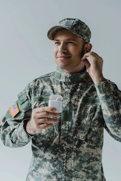 Весёлый солдат в форме и кепке держит футляр для наушников и улыбается изолированно на сером — стоковое фото