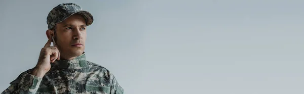Soldado en uniforme y gorra ajustando auriculares inalámbricos aislados en gris, bandera - foto de stock