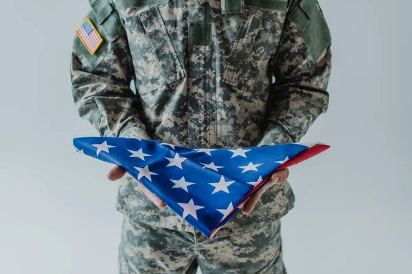 Cropped view of American soldier in uniform holding folded flag of United States during memorial day isolated on grey — Stock Photo