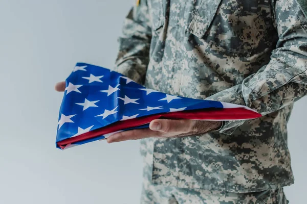 Частичный вид американского солдата в форме, держащего сложенный флаг США в день памяти, изолированный на сером — стоковое фото