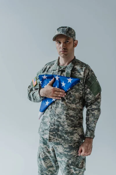 Soldado de boa aparência em uniforme militar segurando bandeira dobrada dos Estados Unidos durante o dia comemorativo isolado em cinza — Fotografia de Stock