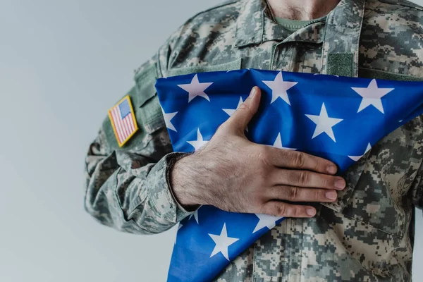 Vista parcial del militar americano sosteniendo la bandera plegada de Estados Unidos durante el día conmemorativo aislado en gris - foto de stock