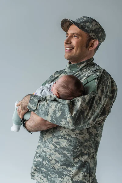 Cheerful father in military uniform and cap hugging newborn boy isolated on grey — Stock Photo