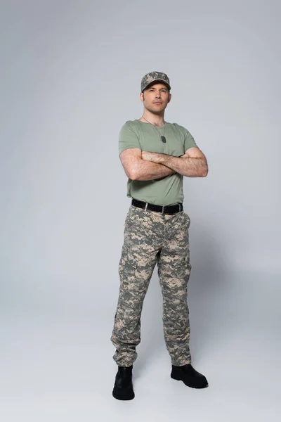 Повна довжина солдата в футболці і кепці, що стоїть зі складеними руками під час пам'ятного дня на сірому — стокове фото