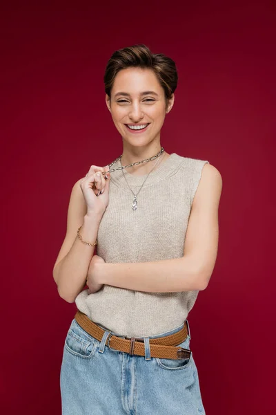 Hübsche junge Frau mit kurzen Haaren lächelt und zieht Halskette auf dunkelrotem Hintergrund — Stockfoto