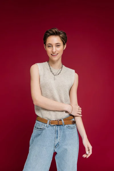 Overjoyed young woman with short hair posing in jeans and sleeveless shirt on dark red background — Stock Photo