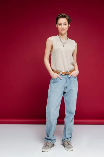 Full length of pleased young woman with short hair posing with hands in pockets of jeans on dark red background — Stock Photo